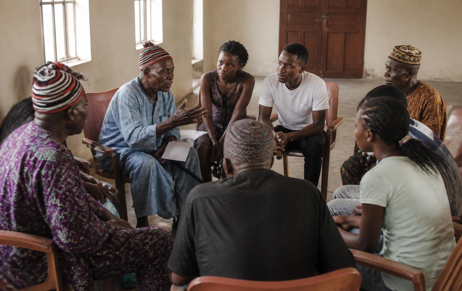 Elders in traditional caps seated in a circle talking with younger members